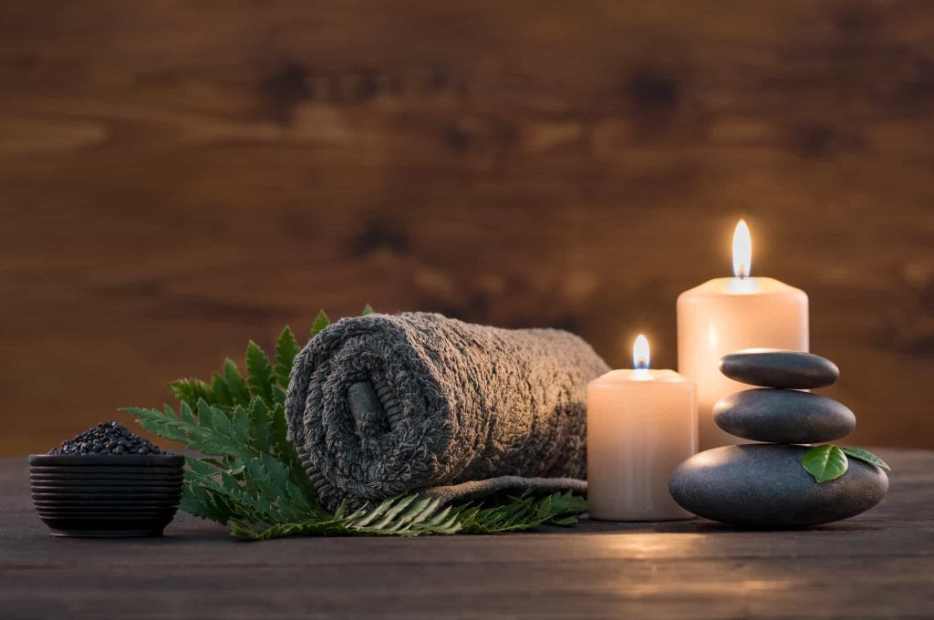 Brown towel on green fern with candles and black hot stone on wooden background.
